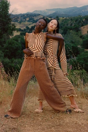 Two women model striped knitwear in an outdoor setting with rolling hills. A darker-skinned woman stands beside a lighter-skinned woman.
