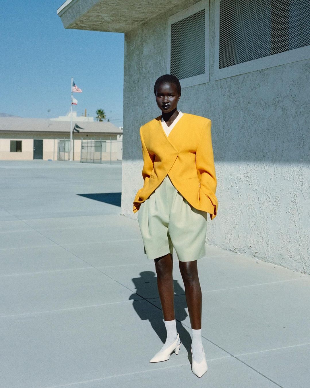 Model wearing a structured yellow blazer and light green shorts standing outdoors against a concrete building under clear sky