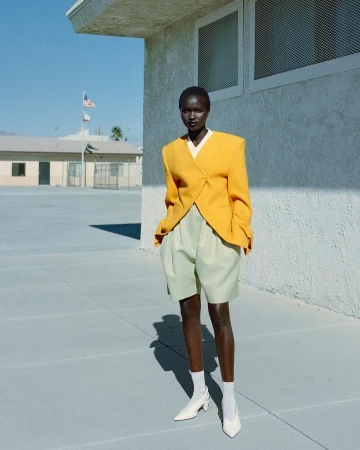 Model wearing a structured yellow blazer and light green shorts standing outdoors against a concrete building under clear sky