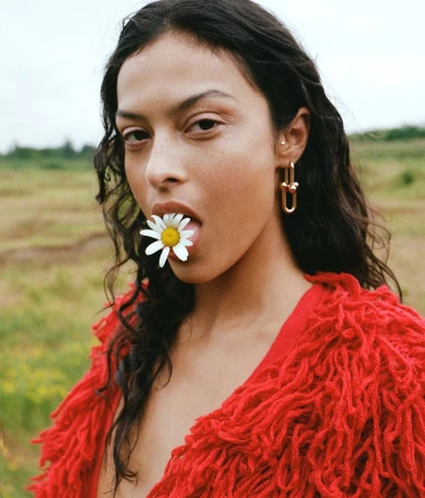 Close-up portrait of a woman with long dark wavy hair wearing a bright red fringed cardigan and holding a white daisy in her mouth.