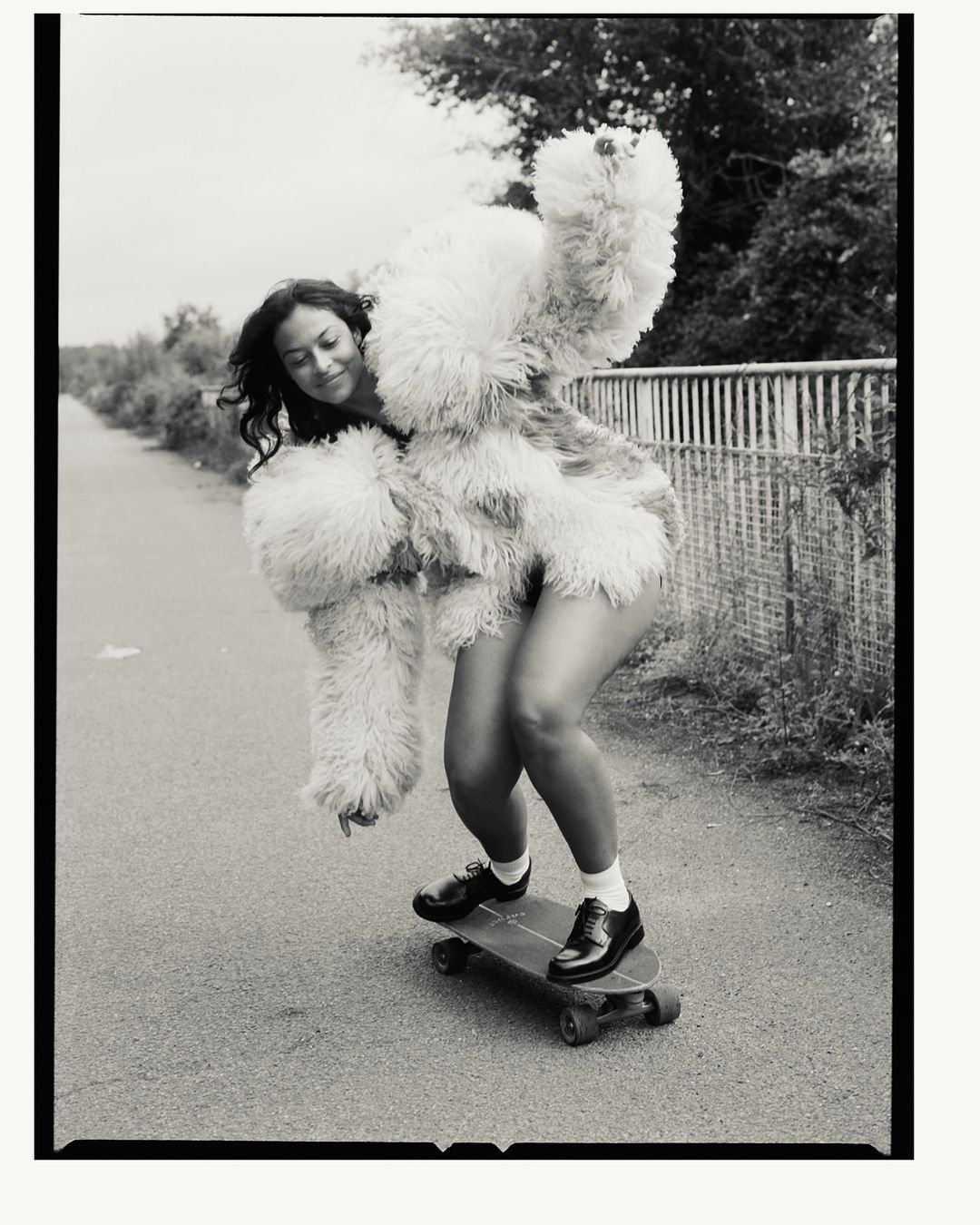 Young woman in a fluffy coat skateboarding on a path with trees in the background. The black-and-white photo captures movement and style.