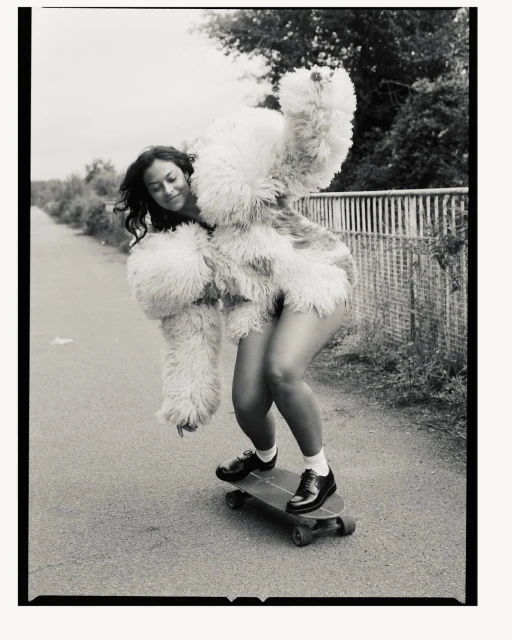 Young woman in a fluffy coat skateboarding on a path with trees in the background. The black-and-white photo captures movement and style.