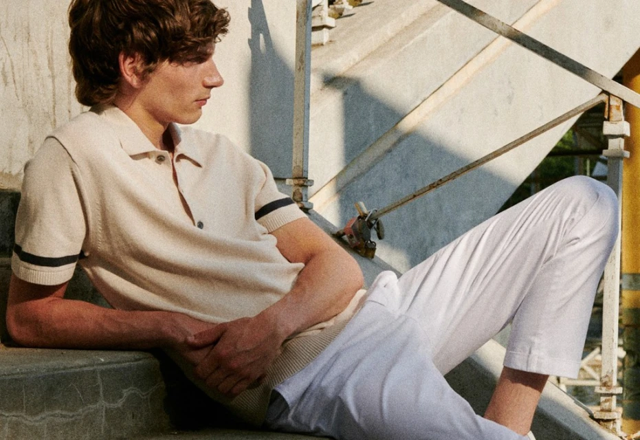 Man in polo shirt and white pants sits on outdoor steps in relaxed pose, bathed in soft light, with architectural backdrop.