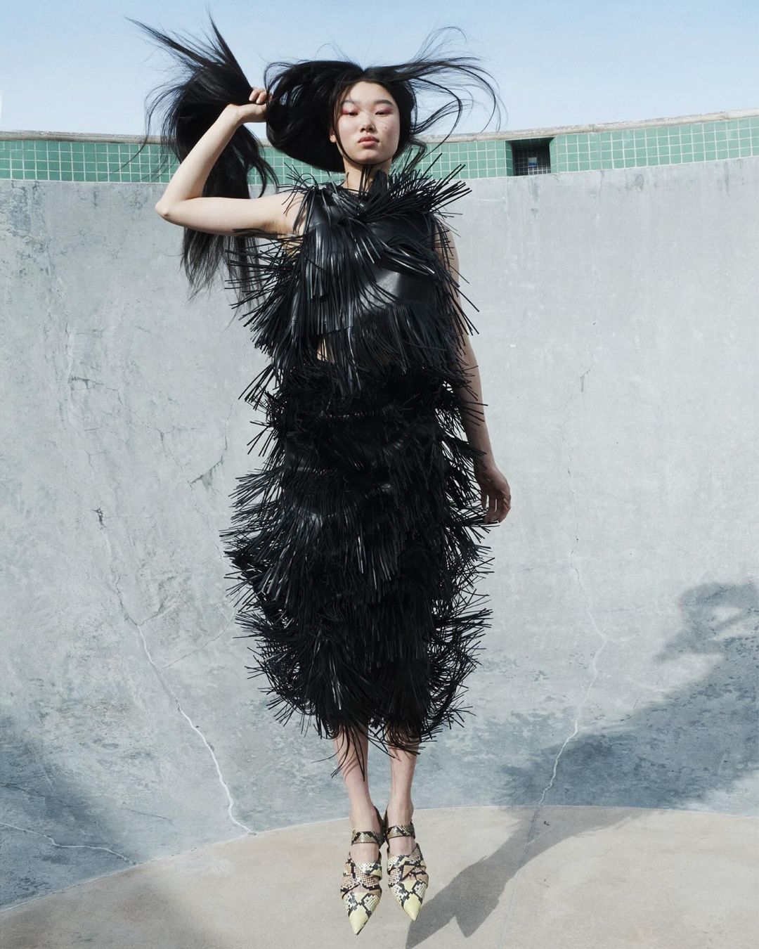 A woman in a fringed black dress jumps against a concrete wall, her long dark hair extending upwards against a bright sky.