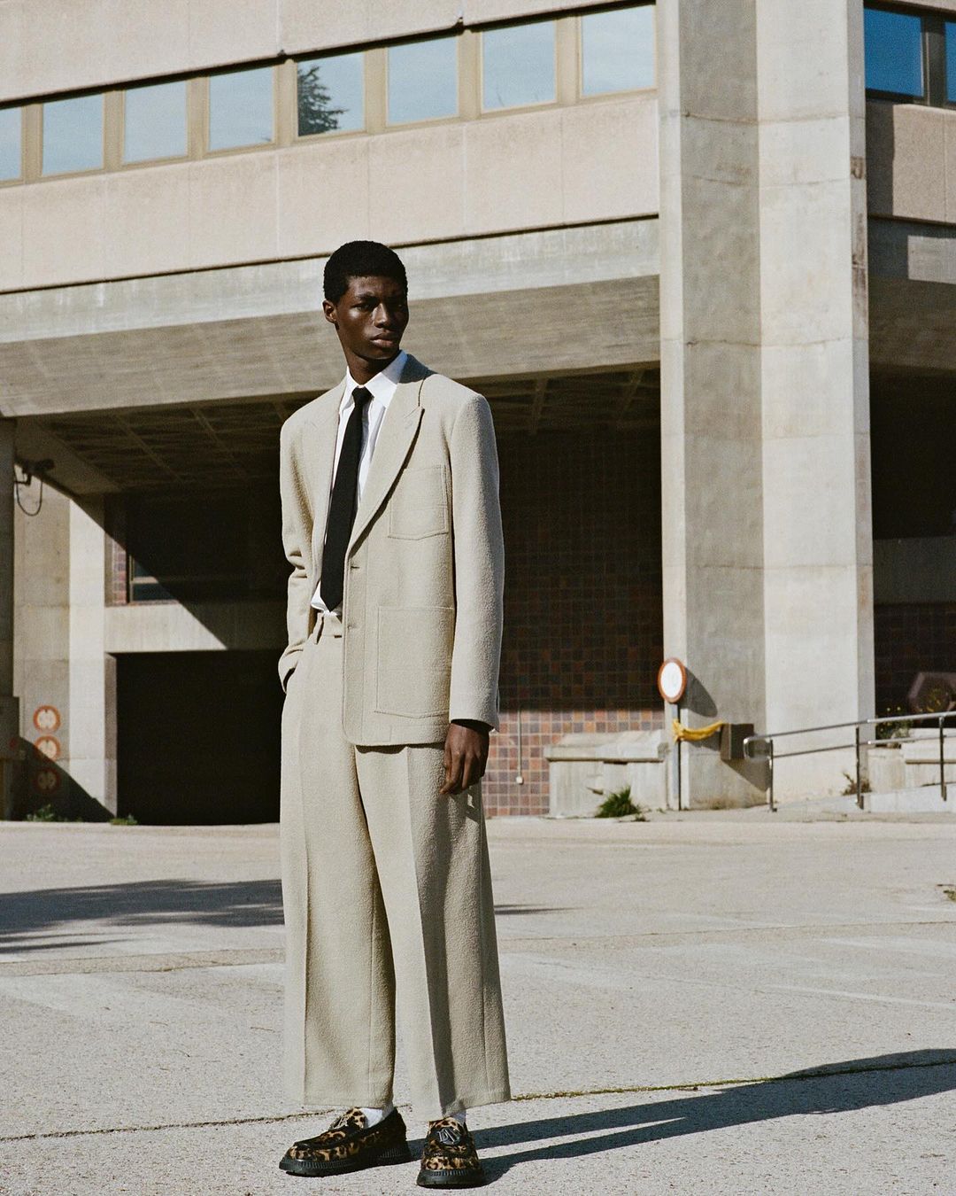 A lone male model stands outdoors in a beige suit with wide-legged pants, a black tie, and patterned shoes, posing against a concrete building.