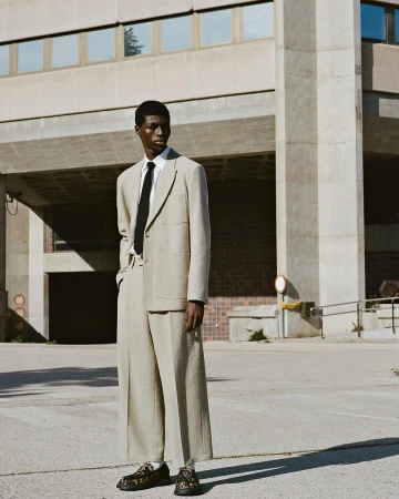 A lone male model stands outdoors in a beige suit with wide-legged pants, a black tie, and patterned shoes, posing against a concrete building.