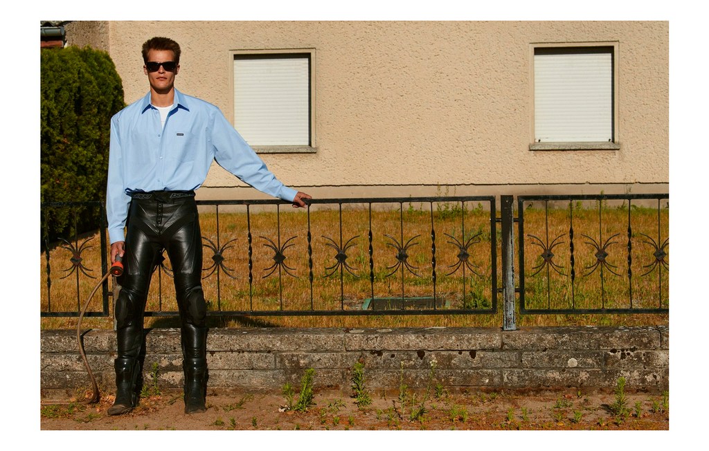 Full shot of an adult man wearing a blue shirt and shiny black leather pants standing outdoors holding a whip near a house.