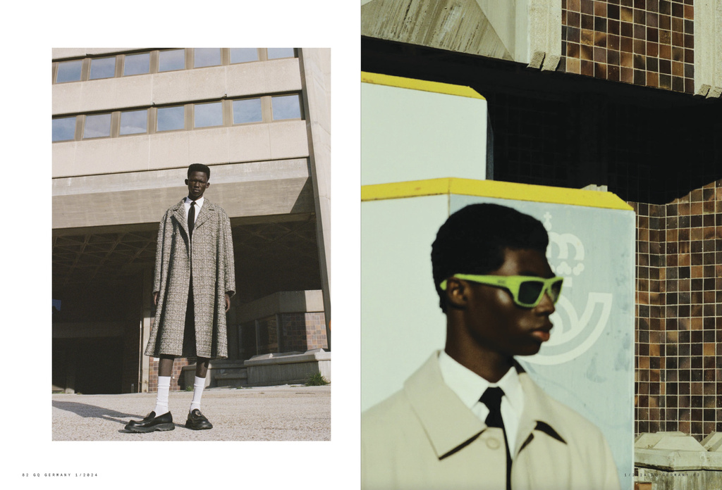 Fashion shot of two Black men in trench coats and suits, one wearing green sunglasses, in an urban setting with brutalist architecture.