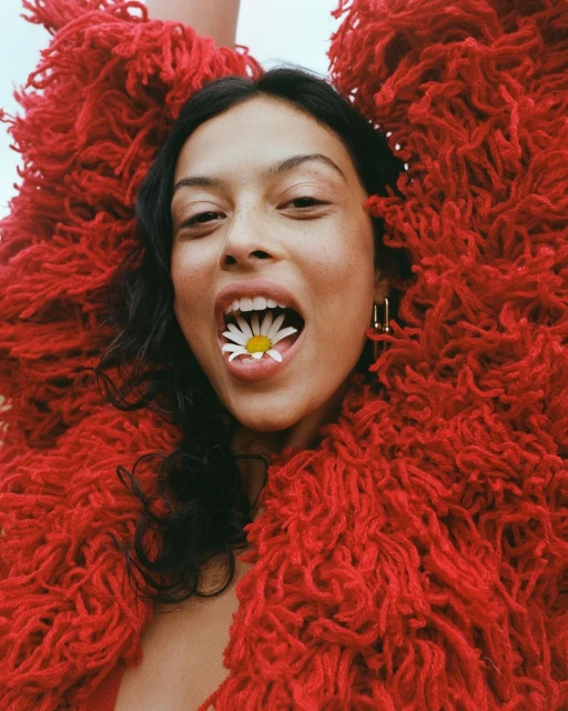 Close-up portrait of a woman with dark hair and red textured clothing, holding a daisy in her mouth, posing indoors with a bright expression.