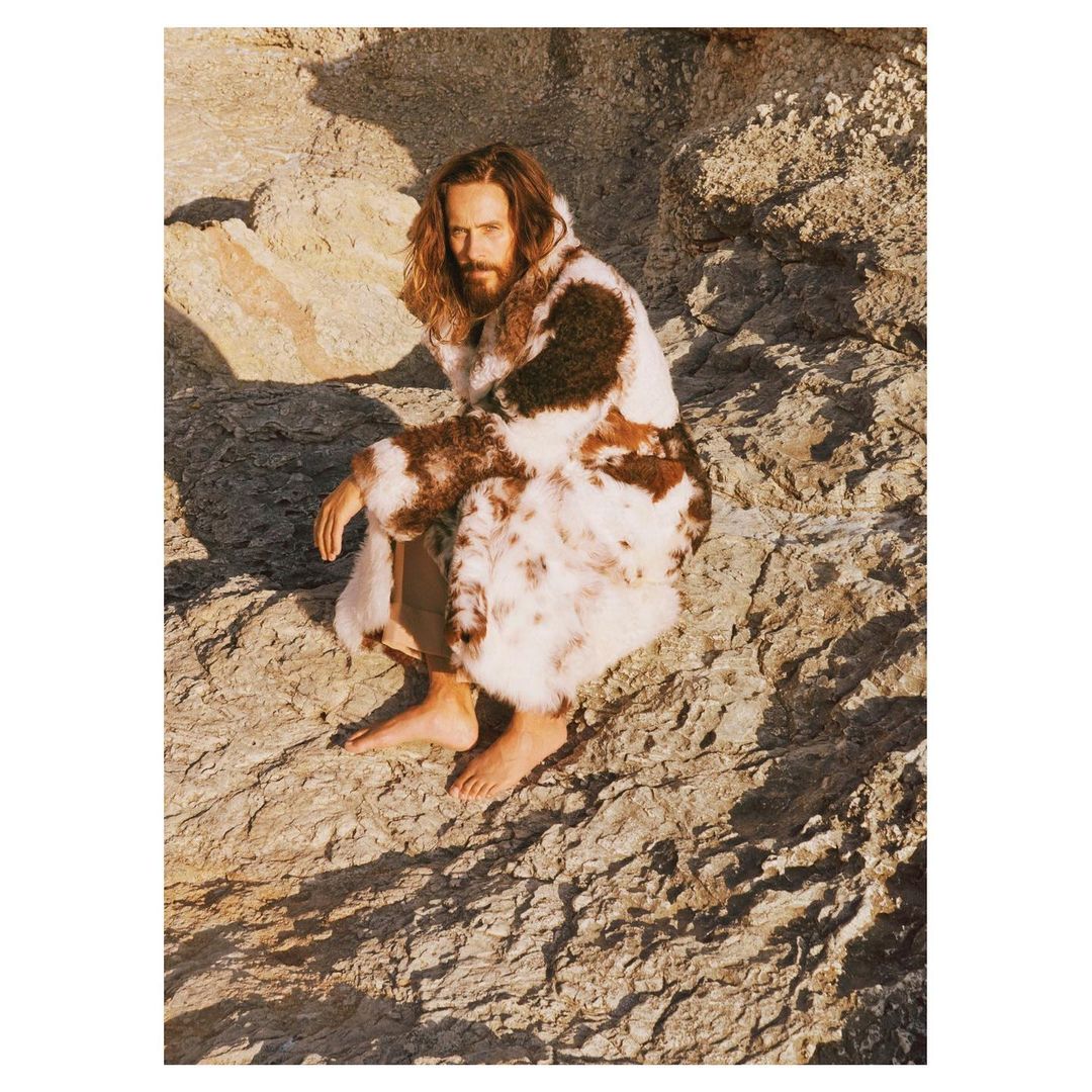 An adult male with long hair and a beard sits barefoot on a rocky surface, wearing a multicolored fur coat in an outdoor setting.