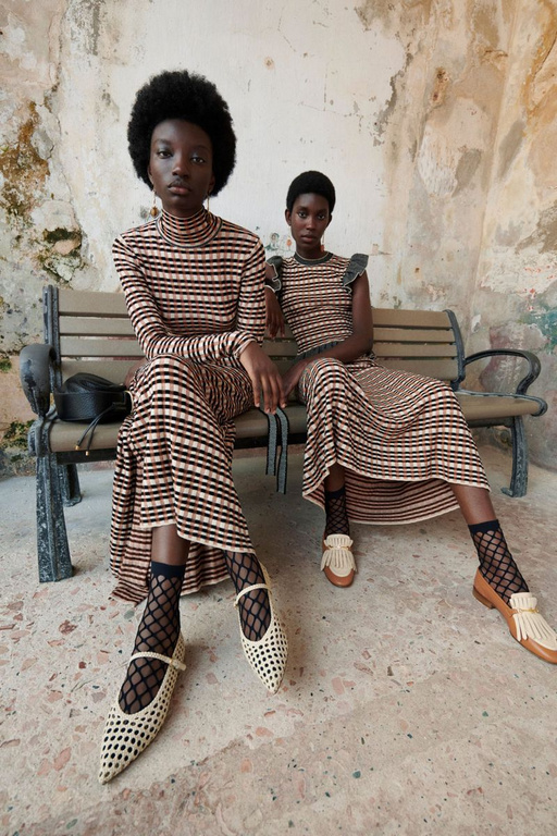 Two Black women in striped dresses sit on a bench, modeling fashion shoes and socks in front of an old textured wall, outdoor fashion shoot.