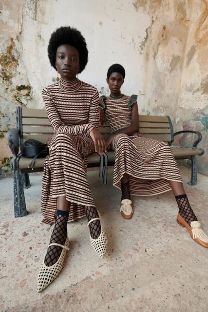 Two Black women in striped dresses sit on a bench, modeling fashion shoes and socks in front of an old textured wall, outdoor fashion shoot.