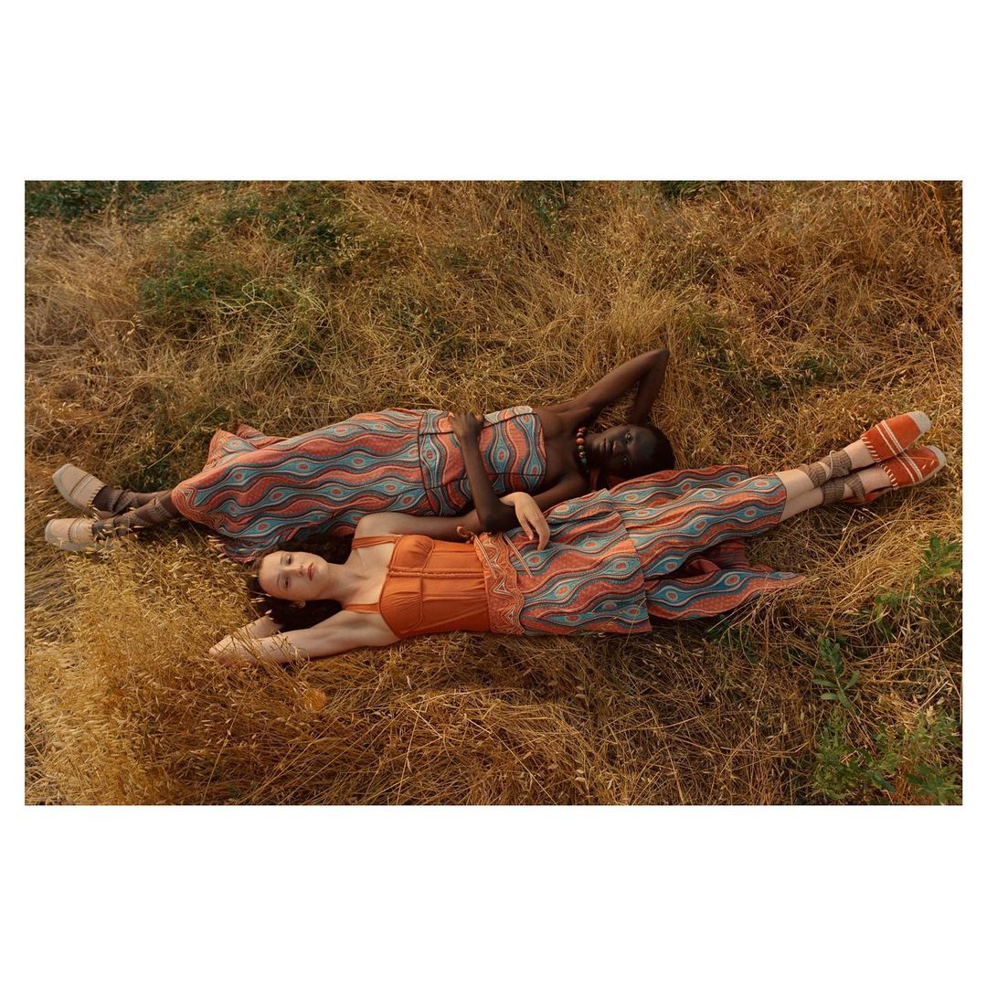 Two women lie in a field of tall grass wearing matching orange patterned dresses, enjoying a serene moment in natural light.