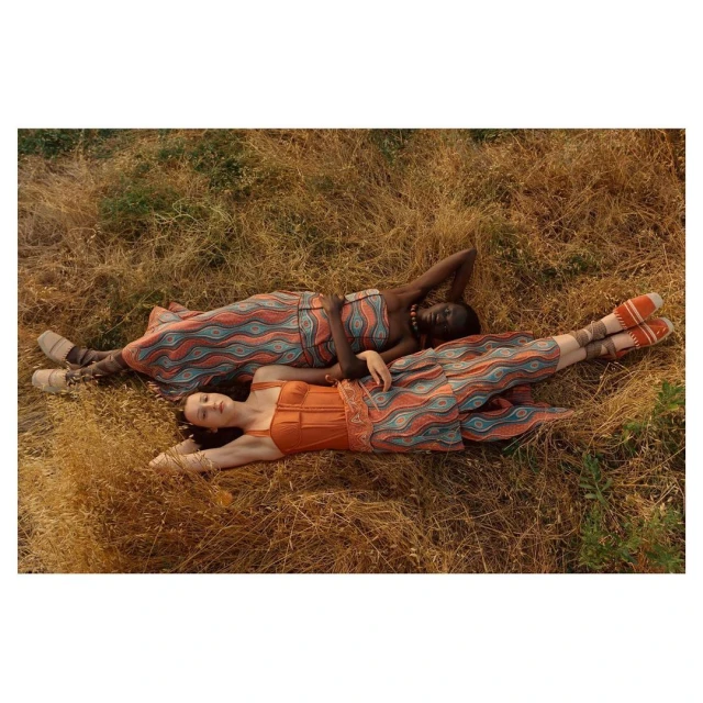 Two women lie in a field of tall grass wearing matching orange patterned dresses, enjoying a serene moment in natural light.