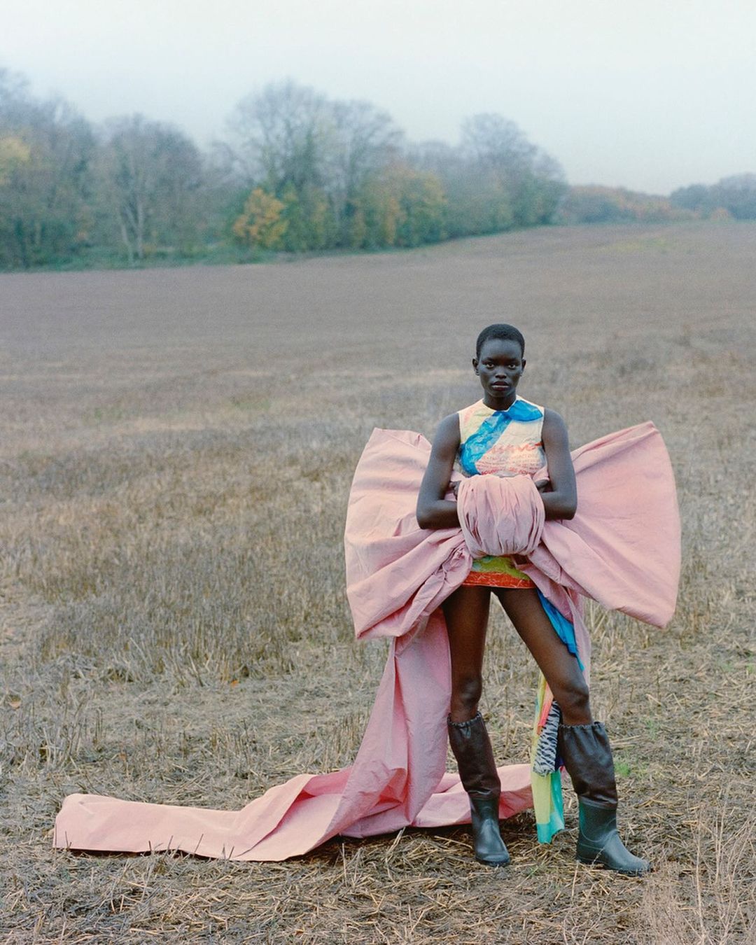 A striking fashion shot features a Black woman in a pale pink bow dress and boots, standing in a field with trees in the background.