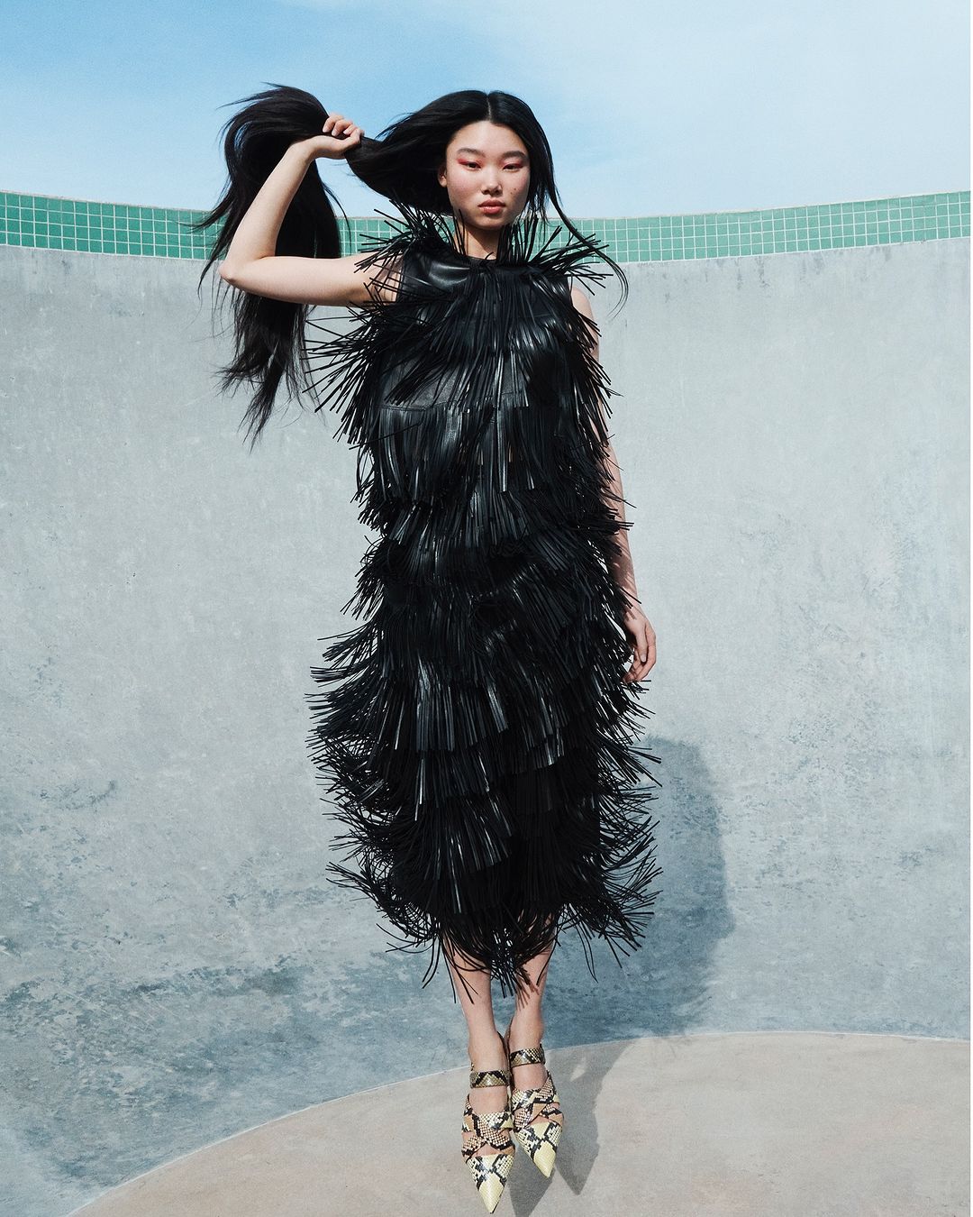 A stylish Asian woman posing mid-air wearing a black, fringed dress and patterned heels in front of a gray wall with a blue sky.