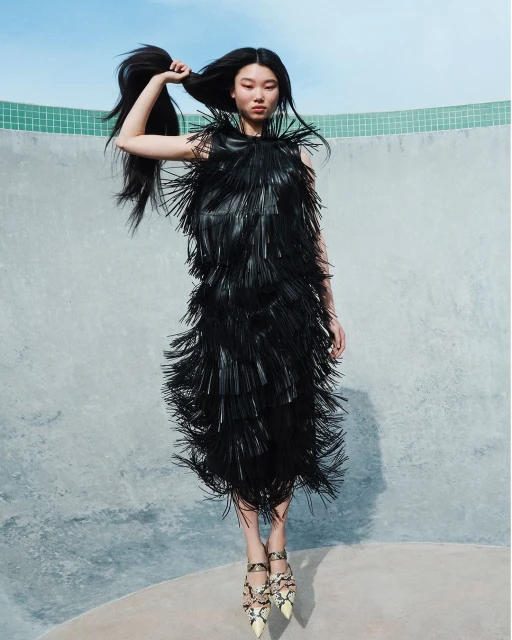 A stylish Asian woman posing mid-air wearing a black, fringed dress and patterned heels in front of a gray wall with a blue sky.