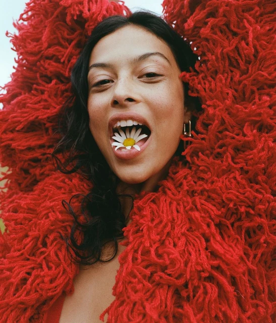 Close-up portrait of a young woman with dark hair and tan skin holding a daisy in her mouth, wearing a red fuzzy coat.