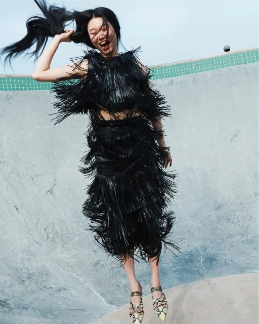 A young Asian woman in a black fringe dress jumps in a concrete setting with a blue sky, showcasing a playful and fashionable moment.