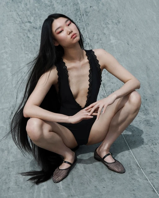 An Asian woman with long, dark hair poses in a black bodysuit and Mary Jane shoes against a gray marble background in a fashion shot.