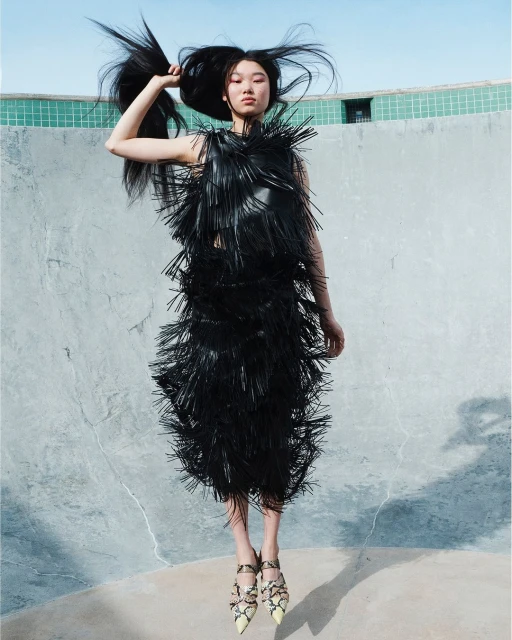 An Asian woman with long black hair wears a black fringe dress and patterned heels, posing in an outdoor setting with a light blue sky.