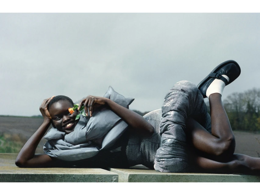 Smiling African female model lies on a stone structure in gray dress, black shoes, and white socks, holding a pink rose in an outdoor setting.