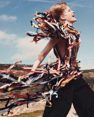 A smiling red-haired model in a fringe-trimmed top and black trousers outdoors against a bright blue sky and rugged hills.