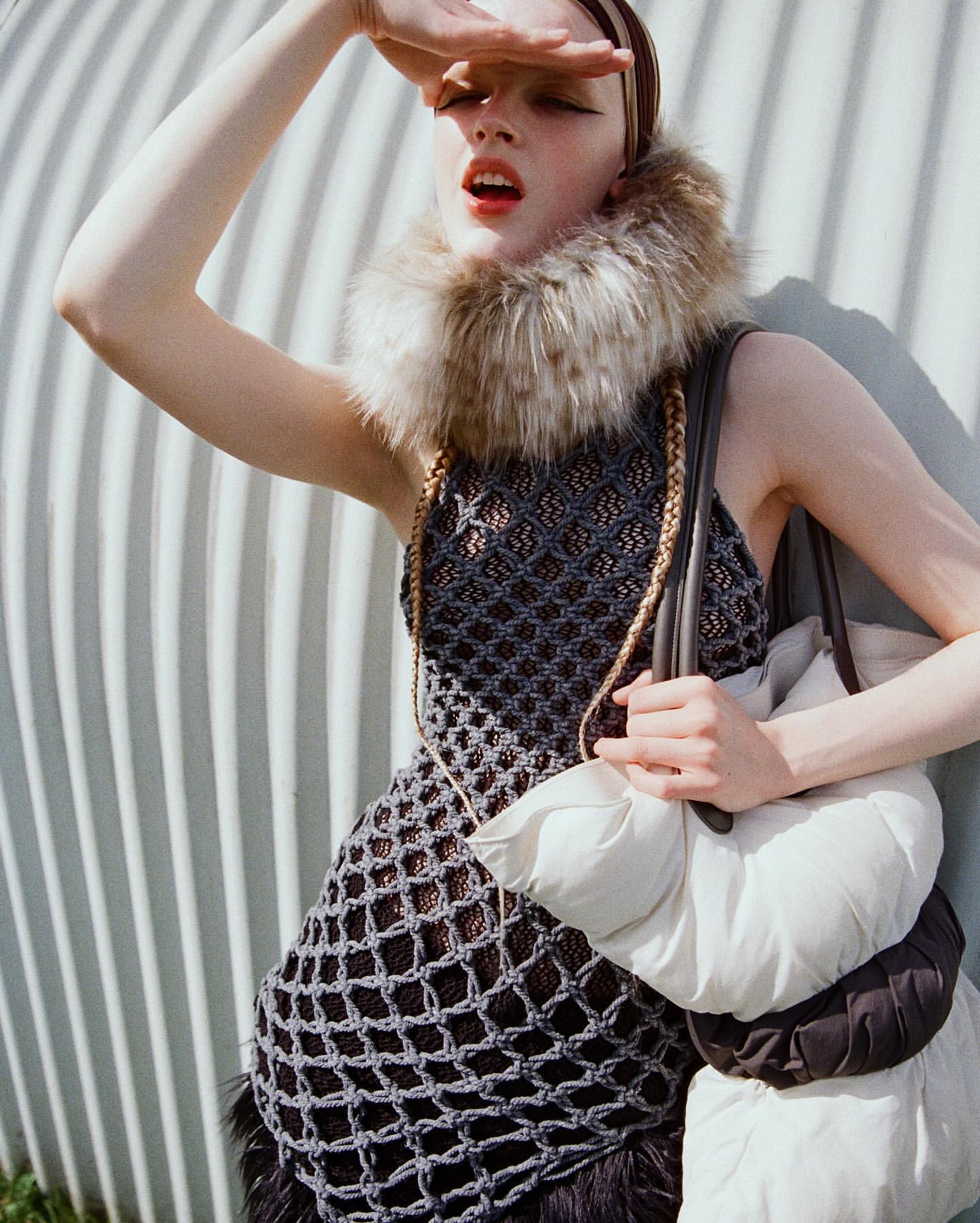 A light-skinned model in fashionable knit dress, fur collar and white bag poses outdoors shielding eyes from the sun with hand.