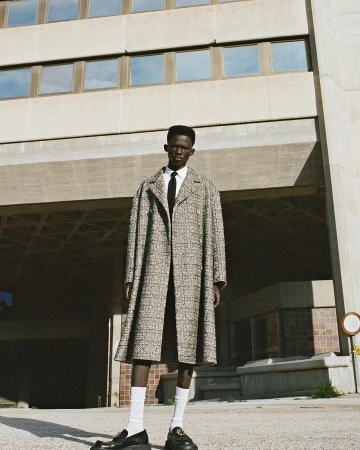 Black man wearing a patterned tweed coat, white shirt, black tie, white socks and black shoes outdoors.