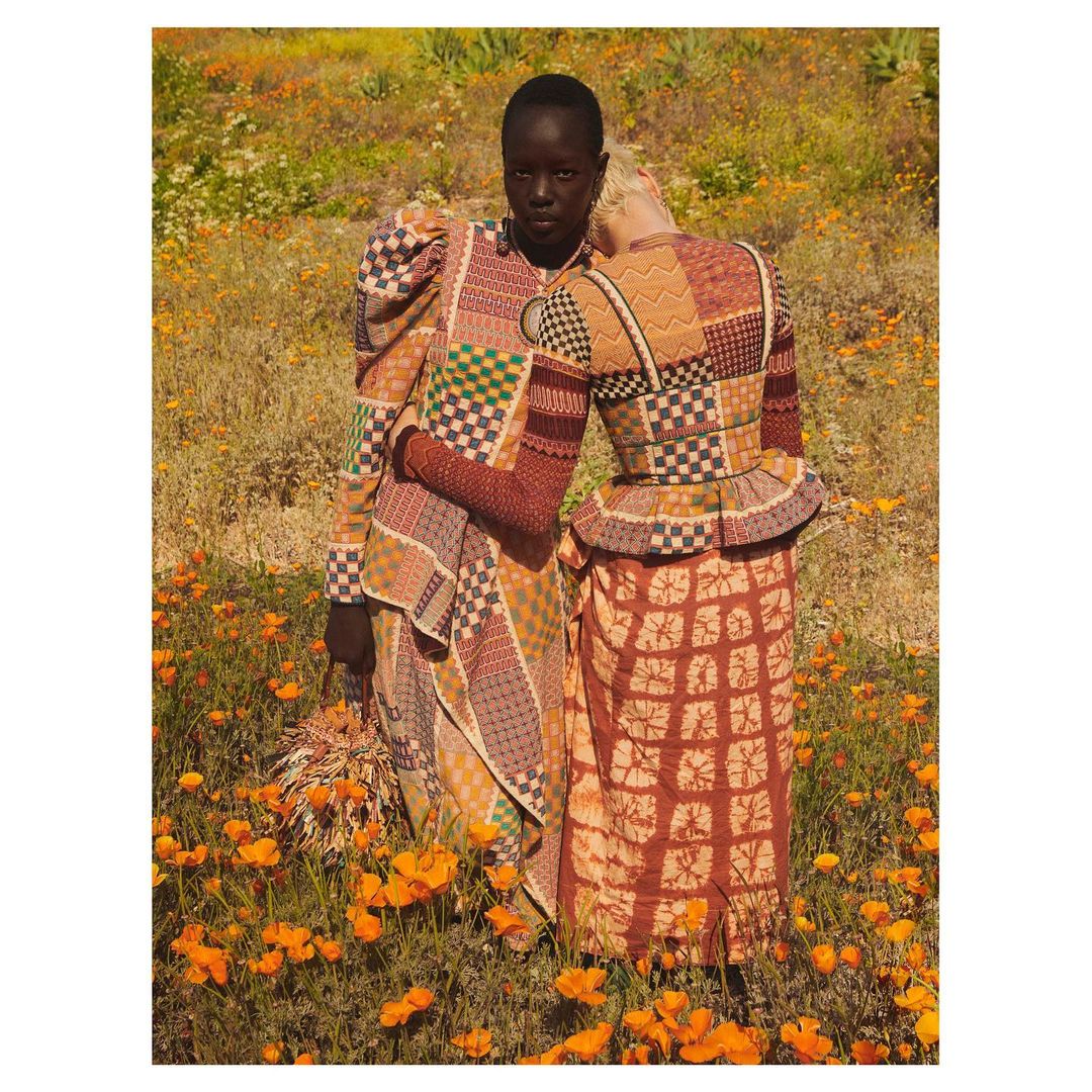 Two women in patterned dresses stand in a field of orange poppies. One rests her head on the other's shoulder in a fashion editorial.