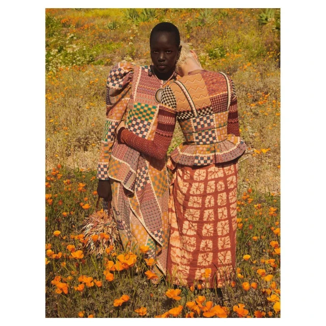 Two women in patterned dresses stand in a field of orange poppies. One rests her head on the other's shoulder in a fashion editorial.