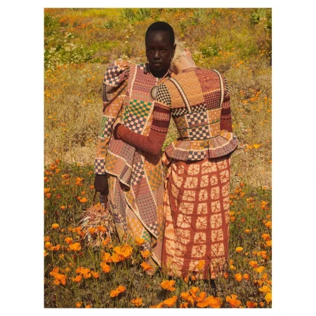 Two women in patterned dresses stand in a field of orange poppies. One rests her head on the other's shoulder in a fashion editorial.