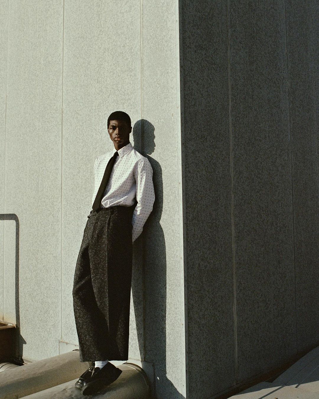 An African American man with a tie and suspenders poses in front of a concrete wall in a studio setting with shadows.