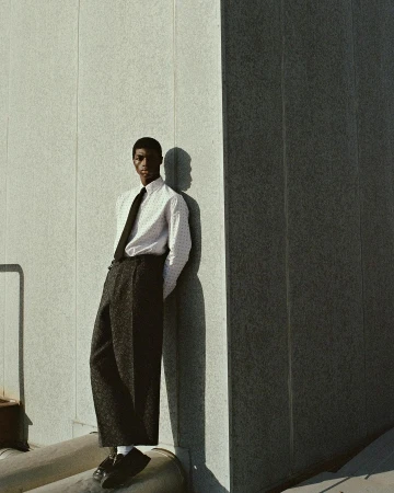 An African American man with a tie and suspenders poses in front of a concrete wall in a studio setting with shadows.