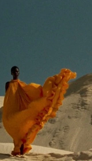 Fashion shot of a black woman walking in the desert in a flowing, bright yellow dress, contrasted against a blue sky backdrop.