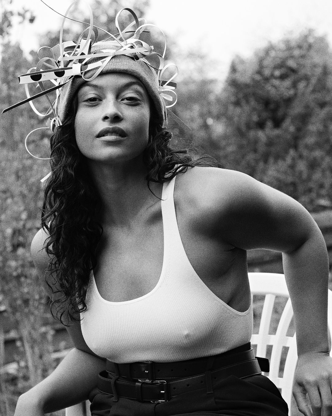 Black and white portrait of a woman wearing a beanie and tank top, accessorized with a decorative headpiece, posing outdoors.