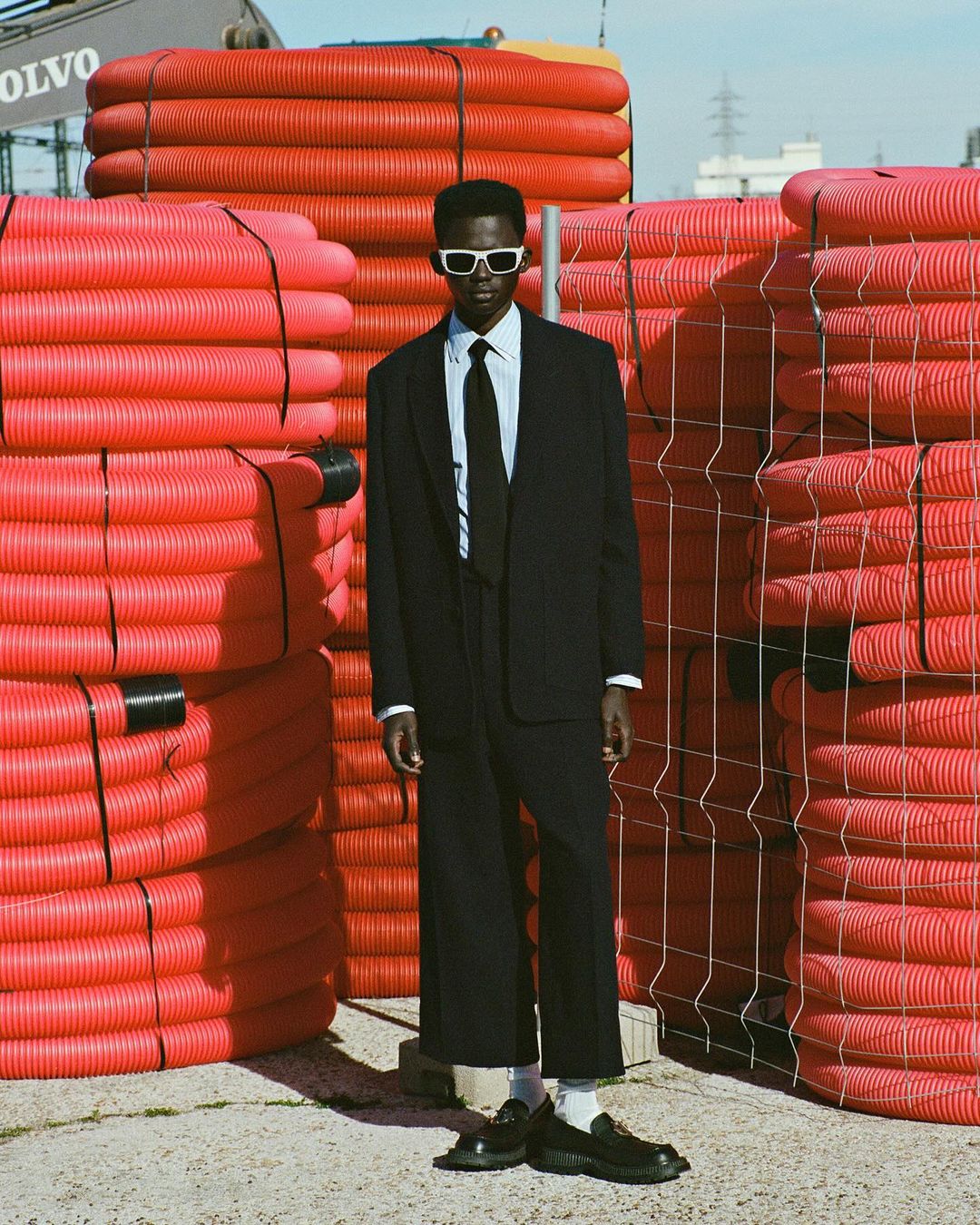 Man in dark suit and sunglasses stands out amid stacks of orange construction pipes under a sunny sky; fashion-forward composition.