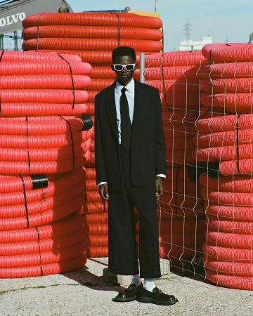 Man in dark suit and sunglasses stands out amid stacks of orange construction pipes under a sunny sky; fashion-forward composition.