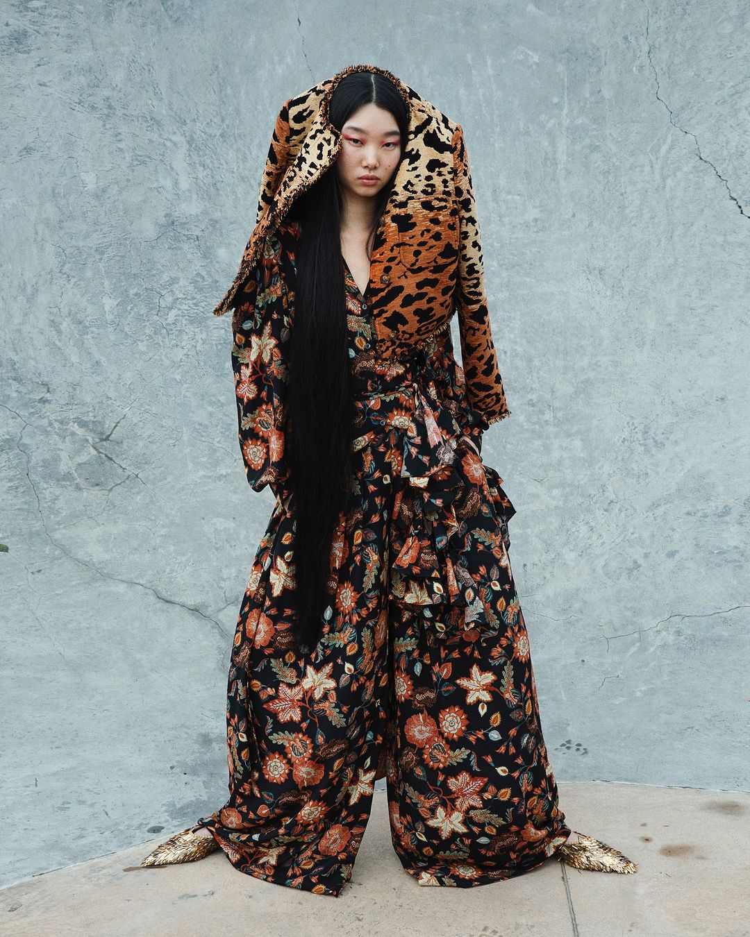 Asian woman in oversized floral pantsuit with leopard print hood poses against textured gray wall, fashion editorial shot in natural light.