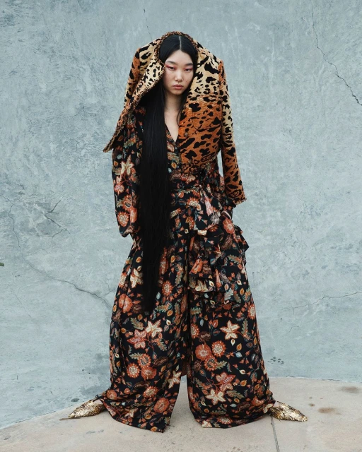 Asian woman in oversized floral pantsuit with leopard print hood poses against textured gray wall, fashion editorial shot in natural light.