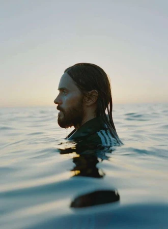 Man in ocean water with dark hair and beard, wearing a dark jacket with white stripes, side profile, horizon, sunset colors.