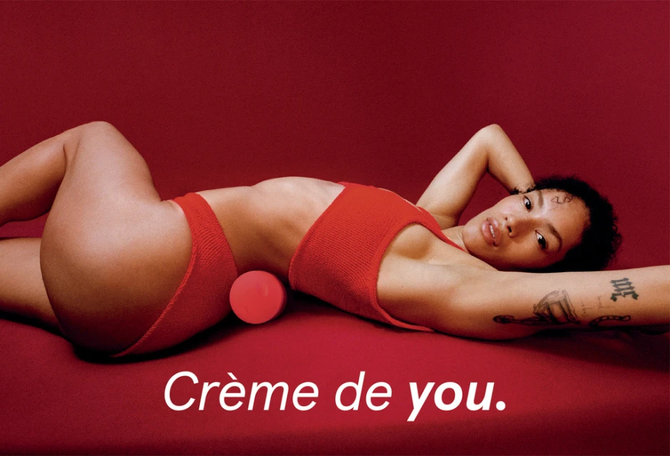 Woman in red lingerie poses reclining against a red background with a red ball; studio lighting and tattoo on her arm visible.