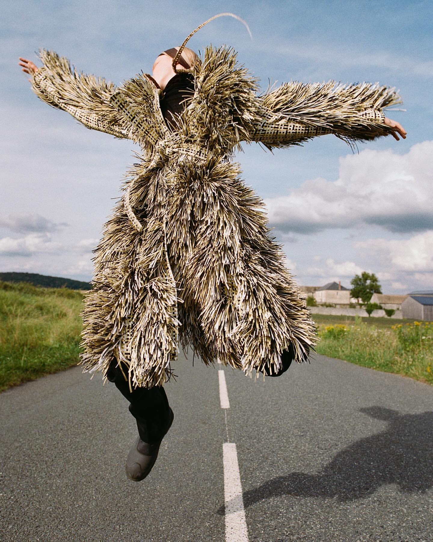 Person in a woven coat jumps on a road, arms outstretched, in a grassy rural landscape under a cloudy blue sky on a sunny day.