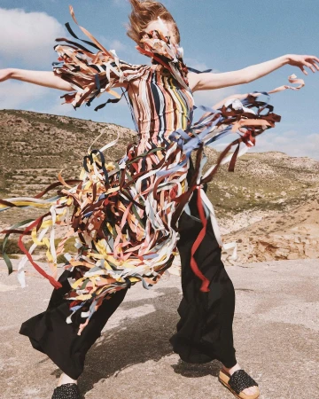 Model in a multi-colored striped top adorned with fluttering ribbons and dark trousers outdoors against a hilly landscape.