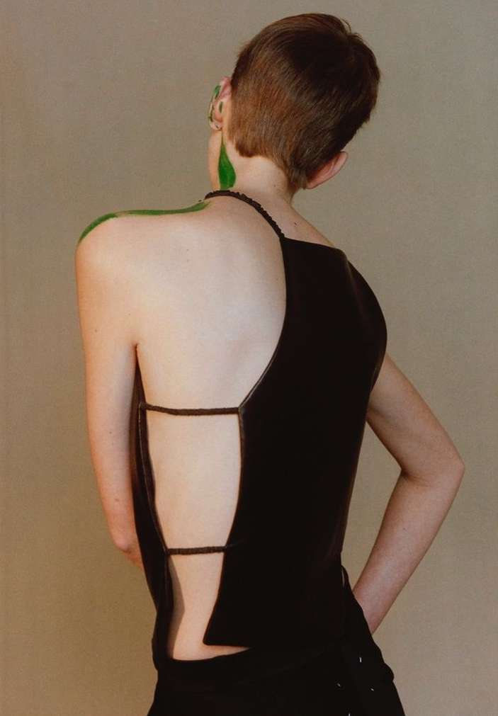 A person with short brown hair and green body paint wears a black, open-back top against a neutral background in a fashion-forward shot.