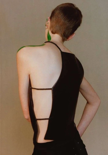 A person with short brown hair and green body paint wears a black, open-back top against a neutral background in a fashion-forward shot.