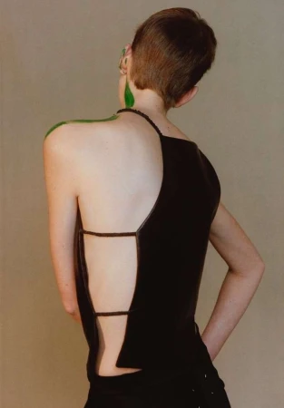 A person with short brown hair and green body paint wears a black, open-back top against a neutral background in a fashion-forward shot.