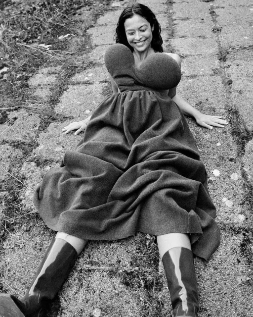 Monochrome portrait of a smiling woman in a sculptural dress and knee-high boots, reclining on a cobblestone surface, natural light.