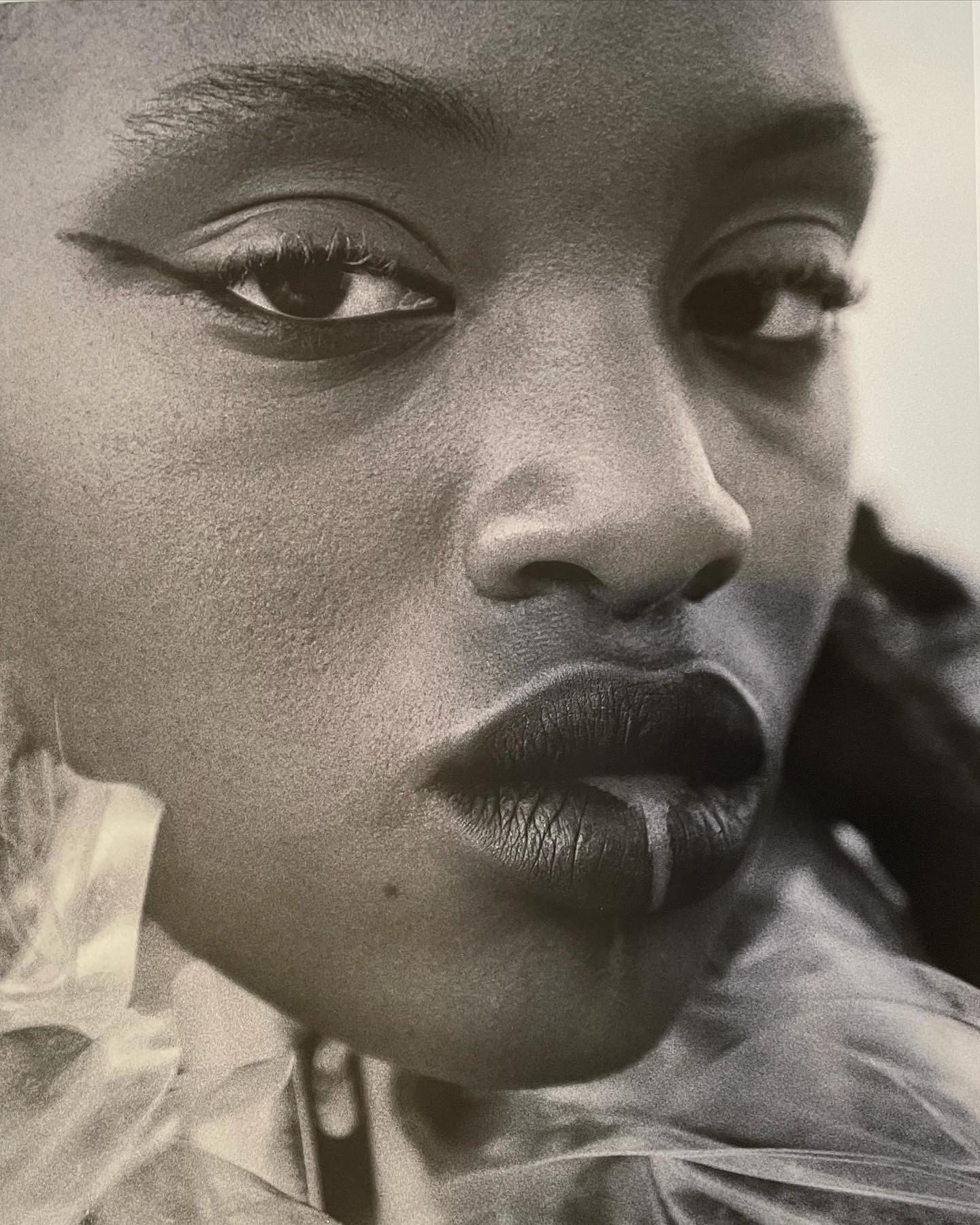 Close-up black and white portrait of a woman with dark hair and winged eyeliner, featuring full lips and a mole on her cheek.