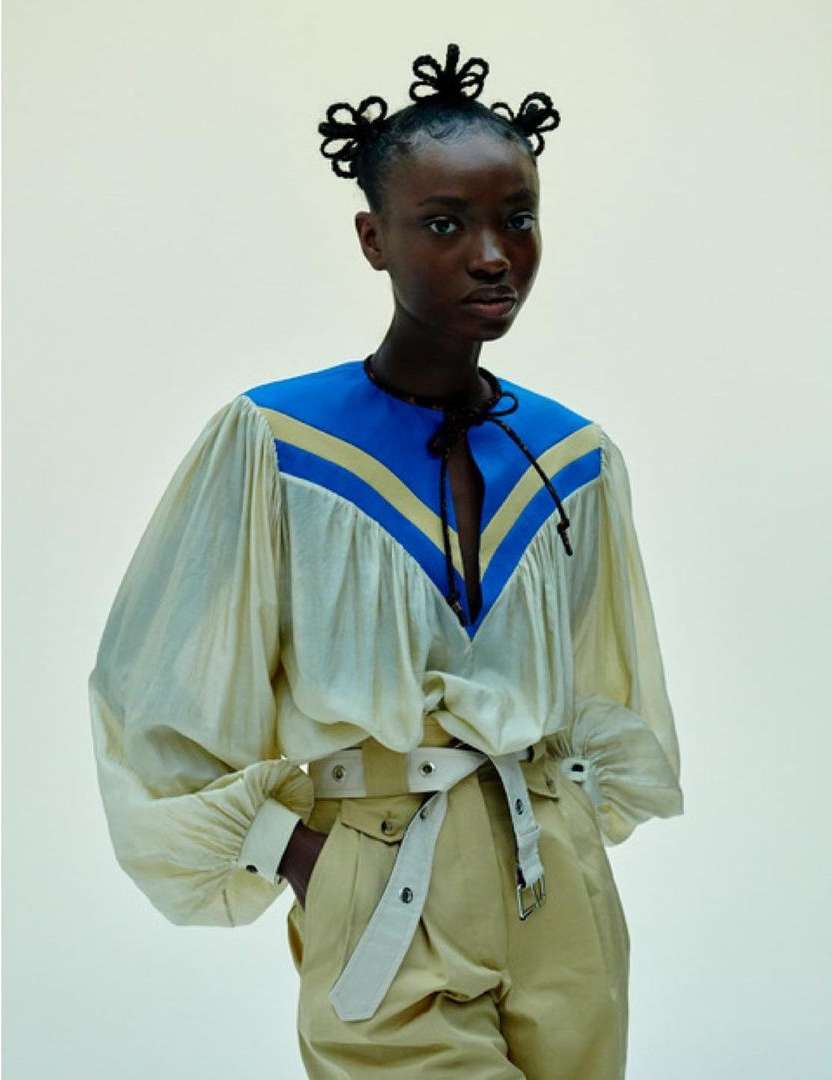 Model with intricate braided hair knots wearing a cream and blue panelled top and khaki belted trousers against a plain wall